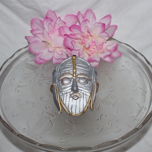 May include: A silver and gold decorative mask with a beard and a detailed design. The mask is sitting on a clear glass plate with a decorative pattern.