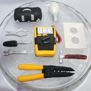May include: A set of fondant tools for a cake decorating project. The tools include a toolbox, a light bulb, a multimeter, a hammer, a screwdriver, a pair of pliers, and a wall outlet.