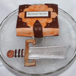 May include: A brown and orange cake decorated to look like a book with the words "Game Level: Married" on the front. A silver sword is on a white plate next to the cake.