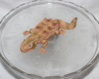 May include: A light brown and green lizard cake topper with a textured surface. The lizard is posed on its belly with its legs outstretched.