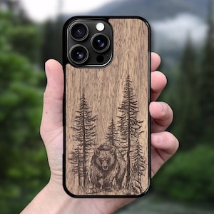 Può includere: Custodia per telefono in legno con bordo nero. Presenta un'incisione dettagliata di un orso in una scena forestale con pini. Il legno ha una venatura naturale e un tono marrone caldo, creando un'estetica rustica.