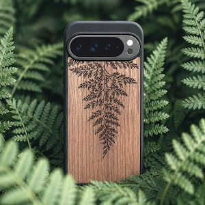 May include: A black phone case with a wooden back featuring a detailed fern design. The case has a camera cutout and is surrounded by green fern leaves. The wood has a natural grain and a warm brown color.