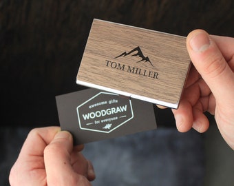 May include: A wooden business card holder with a silver trim. The holder has a mountain design and the name "TOM MILLER" engraved on the front. A black business card with white text reads "awesome gifts WOODGRAW for everyone".