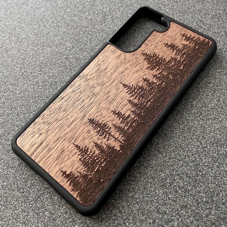 Puede incluir: Una funda rectangular para smartphone con un marco protector negro y un panel trasero de madera plana. La superficie de madera muestra un dise&ntilde;o grabado con l&aacute;ser de un bosque denso con varios pinos de hoja perenne de distintos tama&ntilde;os.
