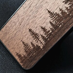 Puede incluir: Una funda rectangular para tel&eacute;fono con una superficie de veta de madera natural y un dise&ntilde;o grabado con l&aacute;ser de un denso bosque de pinos. La madera tiene un tono marr&oacute;n claro, mientras que los &aacute;rboles grabados aparecen en un marr&oacute;n m&aacute;s oscuro.