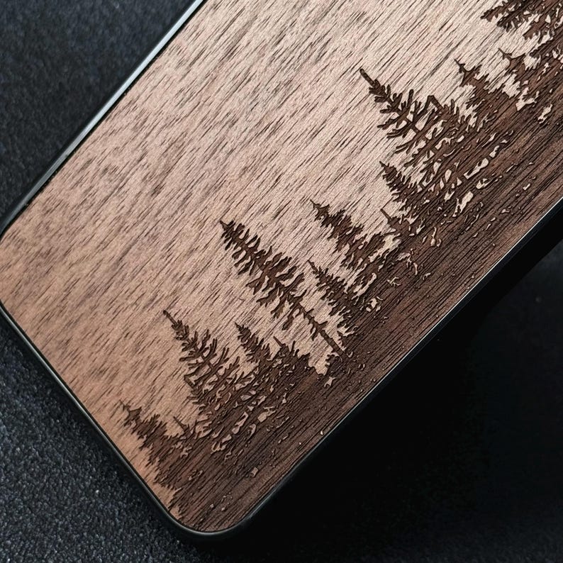 Puede incluir: Una funda rectangular para tel&eacute;fono con una superficie de veta de madera natural y un dise&ntilde;o grabado con l&aacute;ser de un denso bosque de pinos. La madera tiene un tono marr&oacute;n claro, mientras que los &aacute;rboles grabados aparecen en un marr&oacute;n m&aacute;s oscuro.