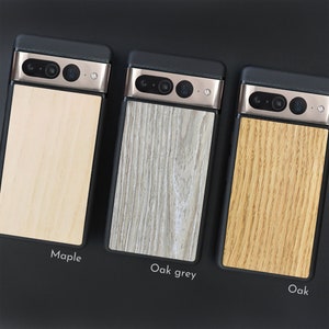 May include: Three phone cases with wooden backings are displayed. The cases have a black frame and a metallic camera bar. The wooden backings are labeled "Maple", "Oak grey", and "Oak", showcasing variations in wood grain and color.