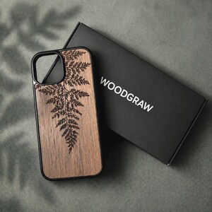 May include: A wooden phone case with a black border and a fern design etched into the wood. The case is next to a black box with the word "WOODGRAW" printed on it. The case is made of wood and has a natural wood grain.