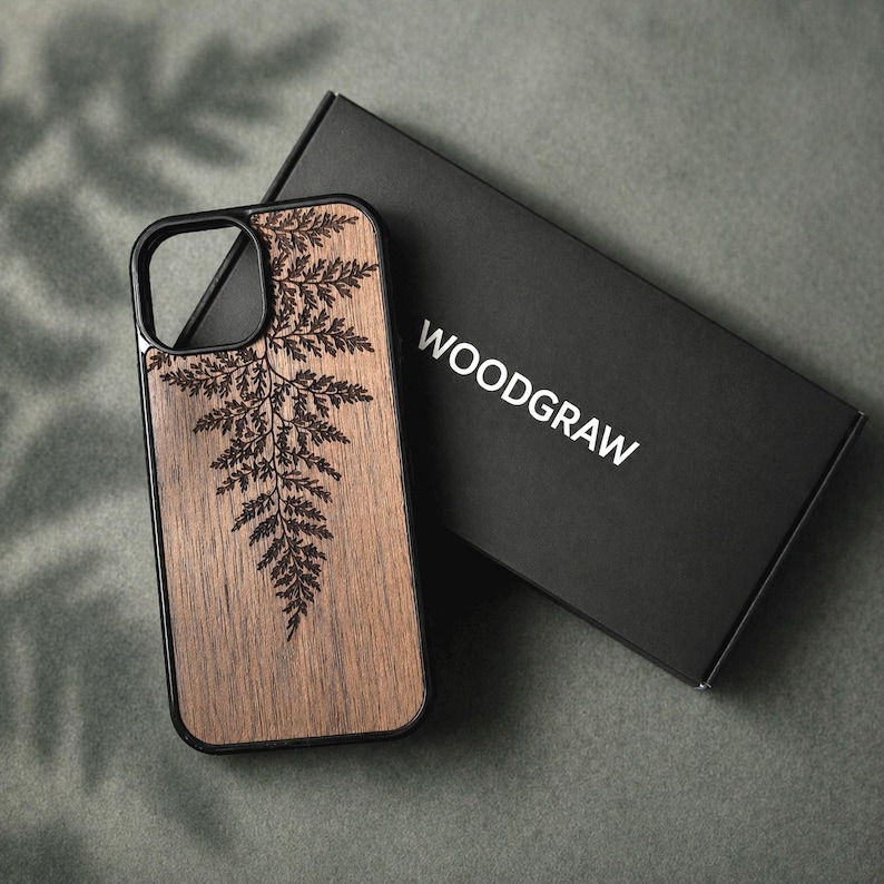 May include: A wooden phone case with a black border and a fern design etched into the wood. The case is next to a black box with the word "WOODGRAW" printed on it. The case is made of wood and has a natural wood grain.