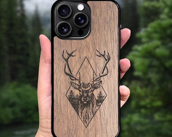 May include: A smartphone case with a wood grain finish featuring a black laser-engraved design of a deer head centered within a geometric diamond shape, with small pine trees depicted at the base of the deer, framed by a black border.