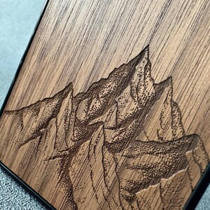 以下が含まれることがあります： レーザー彫刻で山脈が描かれた長方形の木製パネル。垂直に走る自然な茶色の木目と、険しい山の頂や斜面を表現した、質感のある暗い点描の彫刻が対照的です。木材の表面には、山々の起伏が細かく刻み込まれています。
