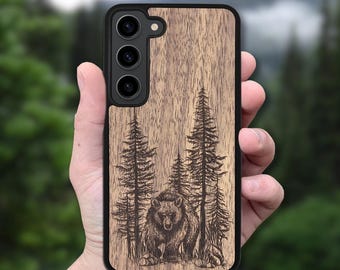 May include: A wooden phone case featuring a detailed engraving of a bear in a forest scene with pine trees. The case has a black border and a natural wood grain finish. The phone case is held in a hand.