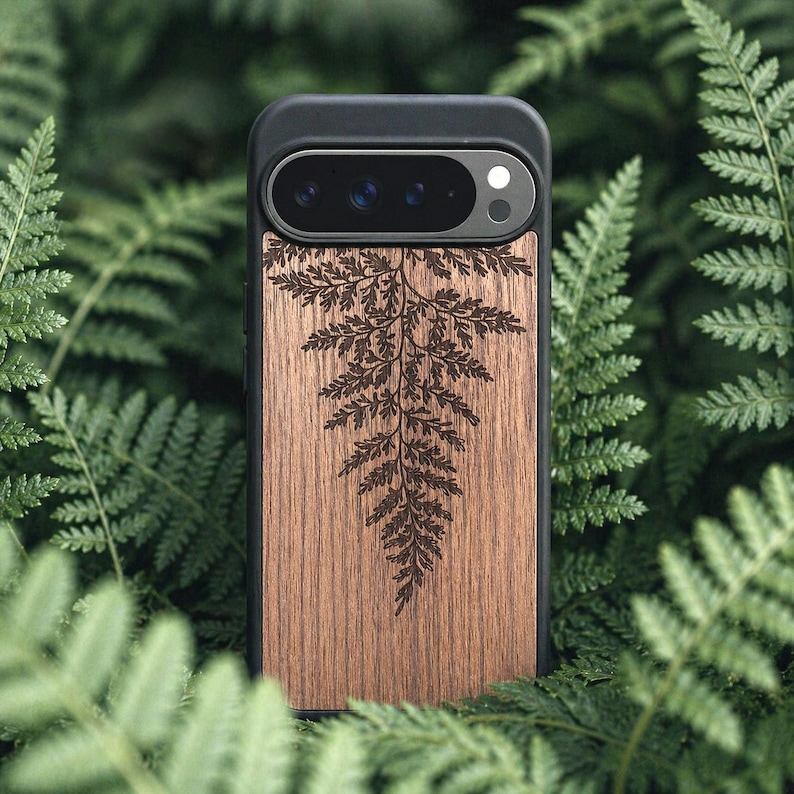 May include: A black phone case with a wooden back featuring a detailed fern design. The case has a camera cutout and is surrounded by green fern leaves. The wood has a natural grain and a warm brown color.