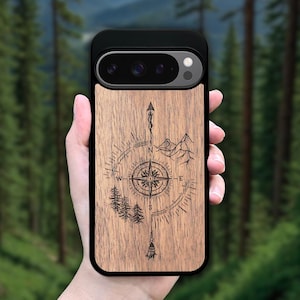 May include: A wooden phone case with a compass, mountains, and trees design. The case has a black border and a camera cutout.