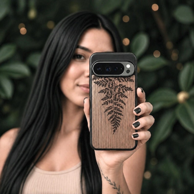 May include: A wooden phone case with a black border and a fern design. The case is held up in front of a person. The fern design is etched into the wood. The phone case has a camera and flash.