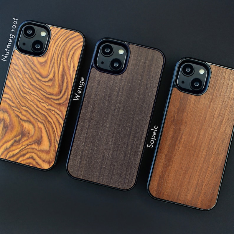 May include: Three wooden phone cases with black trim are displayed against a dark background. The cases feature different wood grains: nutmeg root, wenge, and sapele. The names of the wood types are printed vertically on the side of each case.