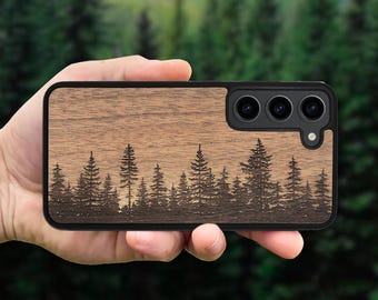 May include: A rectangular smartphone case featuring a natural brown wood grain finish with a laser-engraved silhouette of a dense pine forest across the bottom. The case includes a black frame and a cutout for a triple-lens camera system.