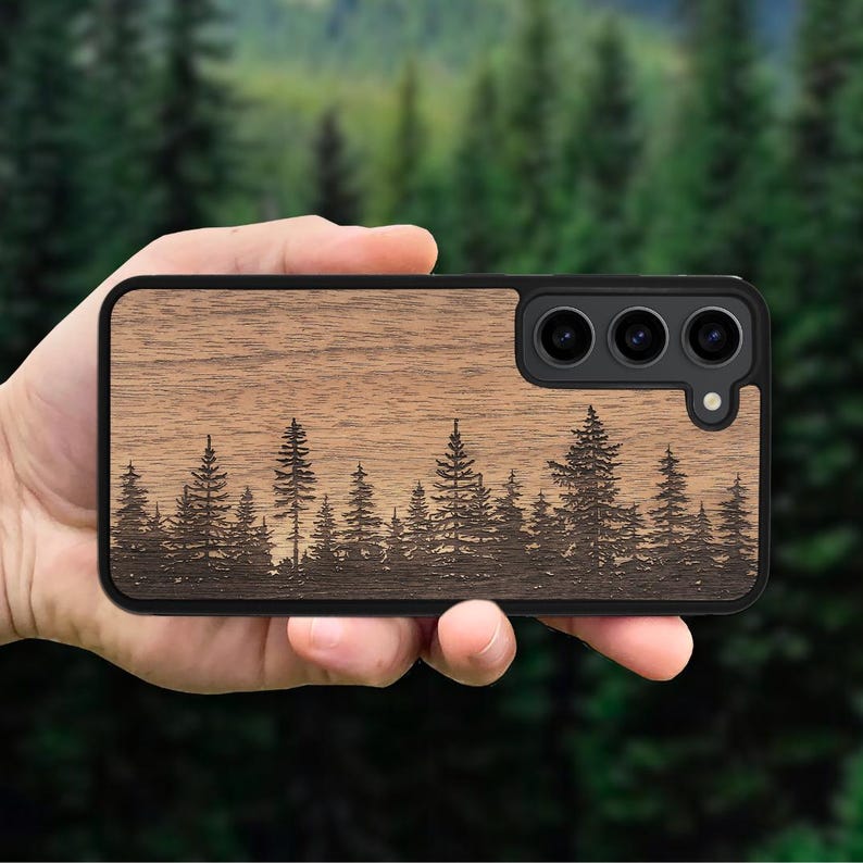 Puede incluir: Una funda rectangular para smartphone con acabado de veta de madera marr&oacute;n natural y una silueta grabada con l&aacute;ser de un denso bosque de pinos en la parte inferior. Incluye un marco negro y un recorte para un sistema de triple c&aacute;mara.