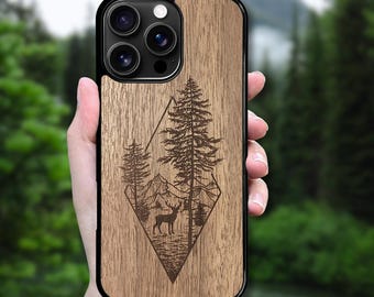 May include: A smartphone case featuring a light brown wood grain finish with a dark brown laser-engraved design. The graphic depicts a deer standing in a forest with pine trees and mountain peaks, all enclosed within a diamond-shaped outline.