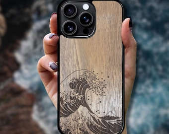 May include: A smartphone case featuring a light brown wood grain finish with a dark, laser-engraved illustration of a large ocean wave. The design includes intricate details of sea foam and a small mountain peak visible at the base of the wave.
