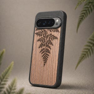 May include: A smartphone case with a wooden panel featuring a fern design. The case has a black border and camera housing. The wood grain is visible, and the fern is etched in a dark brown color. The case is designed to protect the phone.