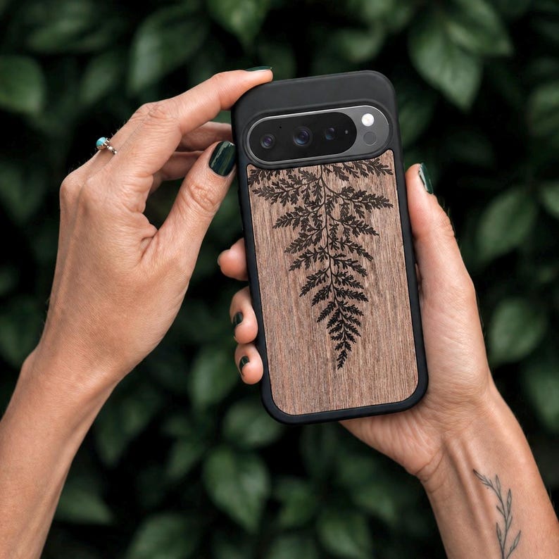 May include: Black phone case with a wooden back featuring a detailed fern design. The case has a black border and a natural wood grain finish. The phone case is held by a person.