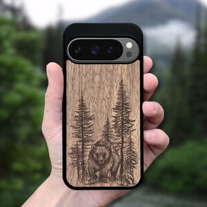 Könnte beinhalten: Schwarze Handyhülle mit Holzmaserungsdesign mit einem eingravierten Bild eines Bären und Kiefern. Die Handyhülle hat einen schwarzen Rand und einen Kameraausschnitt.