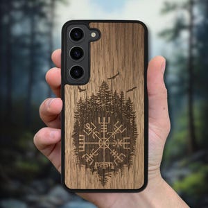 Op de afbeelding: Een houten telefoonhoesje met een zwarte rand. De hoes heeft een gedetailleerd gegraveerd ontwerp van een kompassymbool, een bos en vliegende vogels. Het hout heeft een natuurlijk nerfpatroon en het ontwerp is donkerbruin.