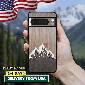 May include: A wooden phone case with a mountain and sun design. The case is made of dark wood with a light wood inlay for the mountain and sun. The case is shown in a hand against a blurry background of trees and mountains.