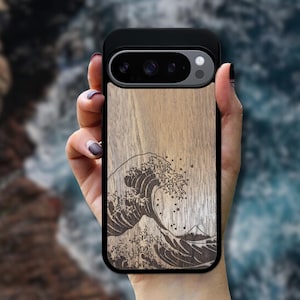 Könnte beinhalten: Eine Handyhülle mit schwarzem Rahmen und Holzrückseite mit einem Wellenmuster. Die Welle ist dunkel gefärbt, das Holz hellbraun. Die Handyhülle wird in einer Hand gehalten.