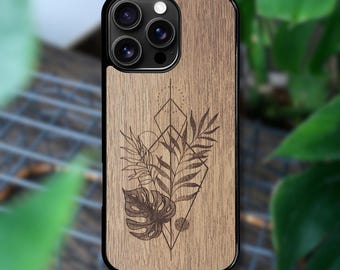 May include: A smartphone case with a light brown wood grain finish featuring a black laser-engraved design of a monstera leaf and fern fronds arranged within a geometric diamond shape, framed by a black border around the camera lens array.