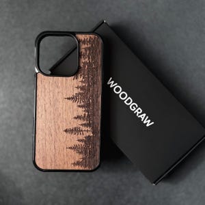 Puede incluir: Una funda de tel&eacute;fono rectangular con un panel trasero de grano de madera marr&oacute;n oscuro que presenta un bosque de pinos grabado con l&aacute;ser. La funda tiene un marco negro y est&aacute; colocada junto a una caja rectangular negra con el texto blanco WOODGRAW.