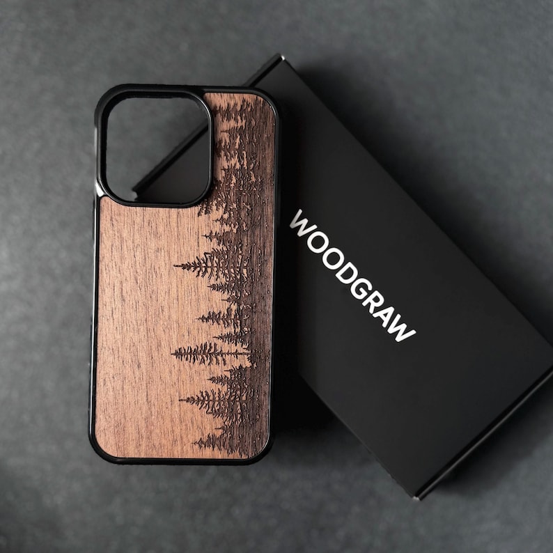 Puede incluir: Una funda de tel&eacute;fono rectangular con un panel trasero de grano de madera marr&oacute;n oscuro que presenta un bosque de pinos grabado con l&aacute;ser. La funda tiene un marco negro y est&aacute; colocada junto a una caja rectangular negra con el texto blanco WOODGRAW.
