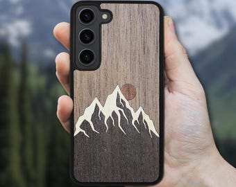 May include: A wooden phone case with a mountain and sun design. The case features a dark wood grain background with a black border. The mountain range is depicted in white, and the sun is a reddish-brown circle.