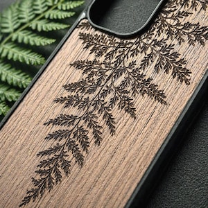 May include: A wooden phone case with a detailed, engraved fern design. The case is made of a light brown wood and features a dark brown fern pattern. The case has a black border and a cutout for the phone's camera.