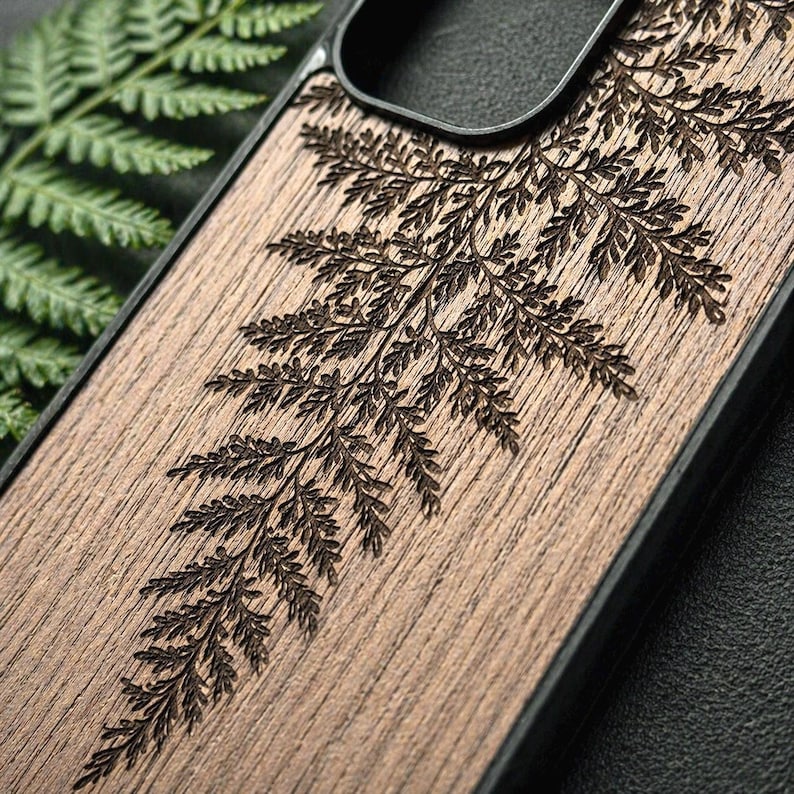 May include: A wooden phone case with a detailed, engraved fern design. The case is made of a light brown wood and features a dark brown fern pattern. The case has a black border and a cutout for the phone's camera.