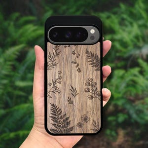 Może przedstawiać: Czarne etui na telefon z drewnianym tyłem z wytrawionymi wzorami botanicznymi. Etui ma czarną obwódkę i naturalny wzór słojów drewna ze szczegółowymi ilustracjami liści i kwiatów.