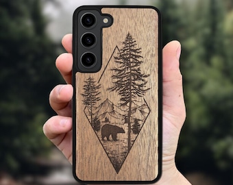 May include: A wooden phone case with a black border. The case features a laser-etched design of a bear, pine trees, and mountains within a diamond shape. The background is a natural wood grain.