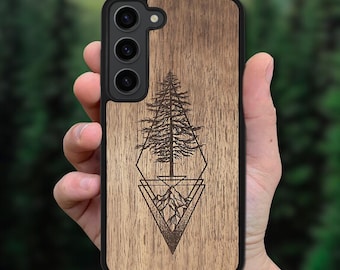 May include: A smartphone case with a wood grain back panel featuring a black laser-engraved design of a pine tree positioned above a mountain range, all enclosed within a geometric diamond shape, set against a black frame with three camera lenses.
