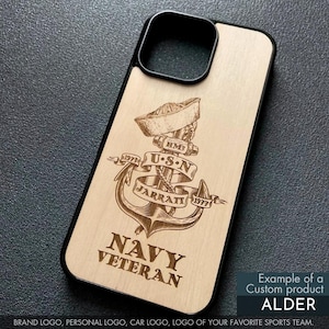 May include: A wooden phone case with a black border, featuring a detailed engraved design. The design includes an anchor, a sailor's hat, and the text "NAVY VETERAN," along with the text "USN" and "JARRATT 1977." The case is labeled "ALDER."