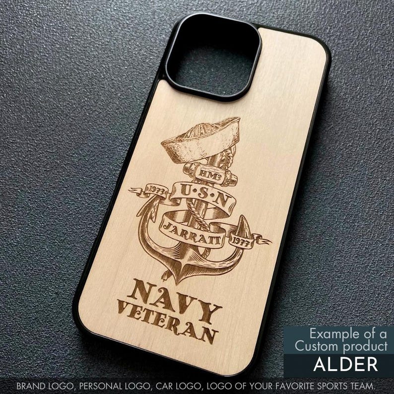 May include: A wooden phone case with a black border, featuring a detailed engraved design. The design includes an anchor, a sailor's hat, and the text "NAVY VETERAN," along with the text "USN" and "JARRATT 1977." The case is labeled "ALDER."