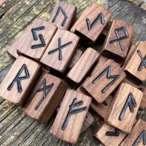 May include: A pile of wooden rune stones, each with a carved black symbol. The rectangular stones are a warm brown colour. The runes, an ancient Germanic alphabet, are used for divination and spiritual practices. The stones are arranged in a random pile.