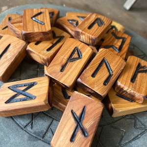 May include: A collection of wooden rune stones with black etched symbols. The rectangular stones are a light brown colour, and the symbols are carved into the wood. The runes are scattered on a grey surface.