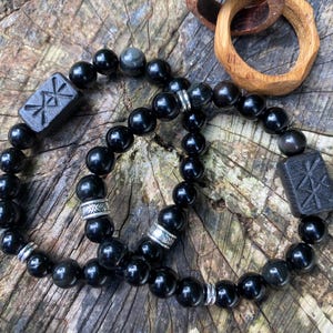 Puede incluir: Dos pulseras de cuentas negras con detalles plateados y dijes de madera con un símbolo tallado. Las pulseras están sobre una superficie de madera.