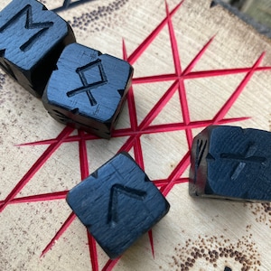 May include: Four black wooden dice with different runes carved into each side. The dice are on a wooden board with red lines and a carved rune in the background.