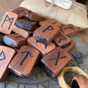 May include: A collection of wooden rune stones with carved black symbols. The rectangular stones are various shades of brown, with some showing darker edges. A wrapped package with a tag is in the background, suggesting a gift or purchase.