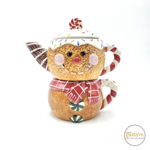 May include: A novelty gingerbread man teapot and cup set. The teapot has a white-iced lid with sprinkles, a red nose, and pink cheeks. The cup has a red and white plaid scarf and a green candy button.