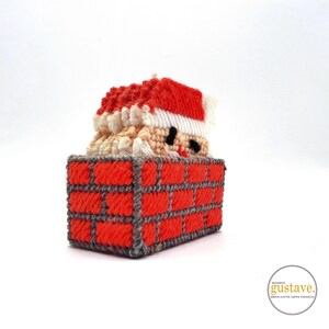 Peut inclure: Ornement de Noël fait à la main représentant le Père Noël sortant d'une cheminée en briques rouges. Le chapeau du Père Noël est rouge avec une bordure blanche, et il a une barbe blanche. La cheminée est en fil rouge avec des joints de mortier gris.