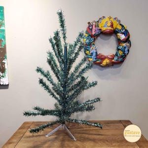 May include: A green tinsel Christmas tree with a silver metal stand. The tree is on a wooden table. A wreath with a nativity scene is on the wall behind the tree.