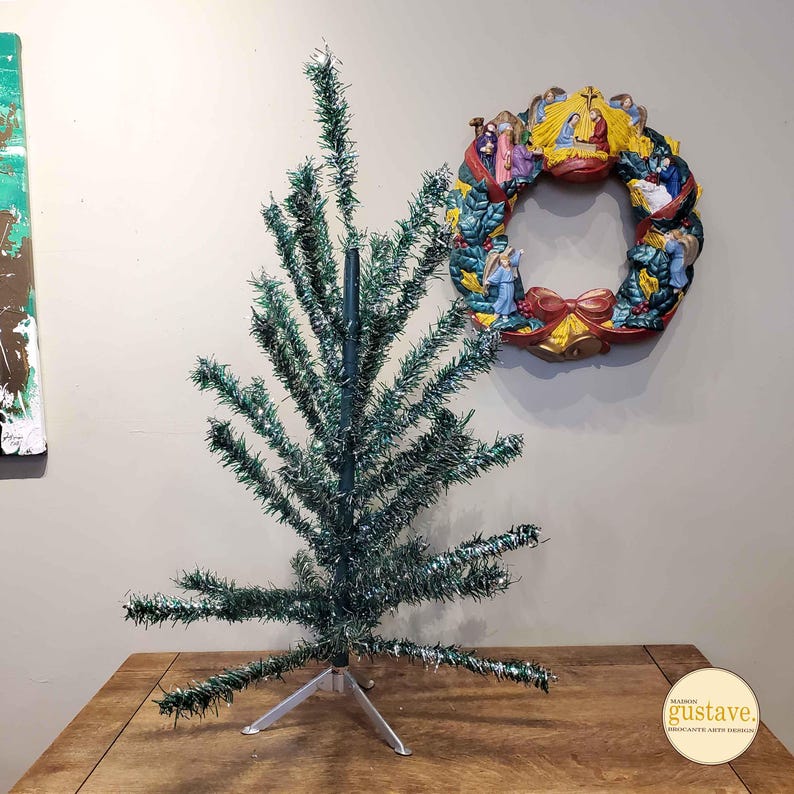 May include: A green tinsel Christmas tree with a silver metal stand. The tree is on a wooden table. A wreath with a nativity scene is on the wall behind the tree.